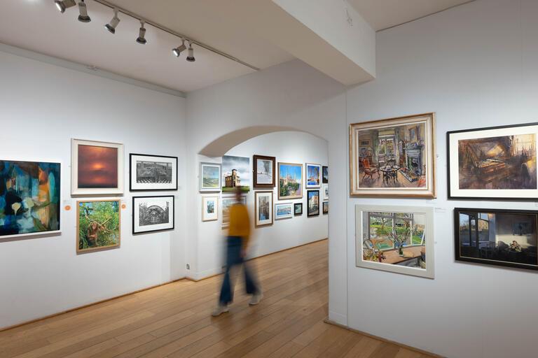 Lady in yellow jumper walking through exhibition with artworks on either side