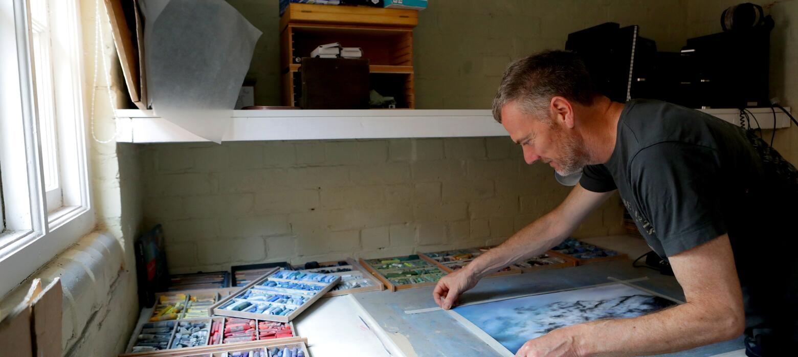 Artist Dave Roberts PS in his studio, taping a pastel drawing to a board.. 