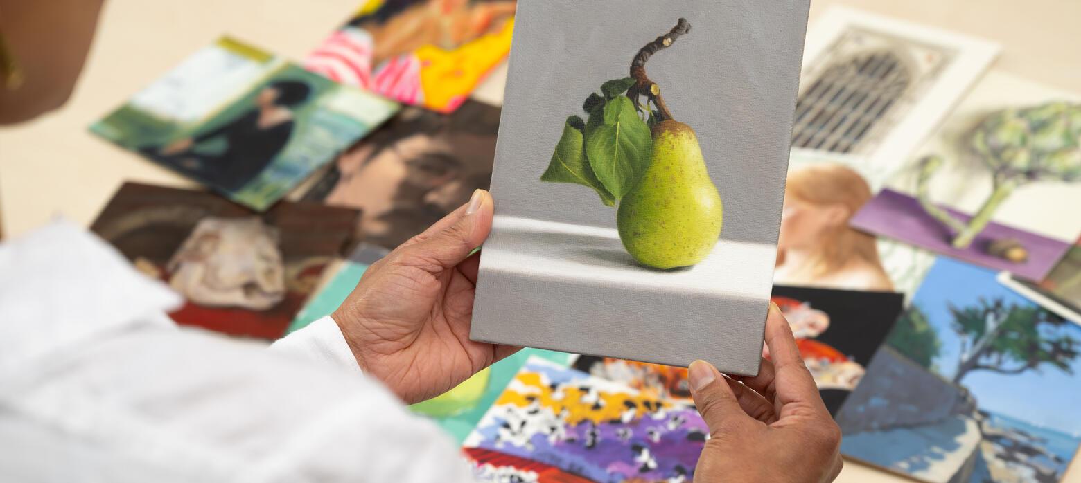 Person holding postcard sized oil painting of a pear with other artworks in the background