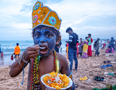 Image of a boy in Indian traditional clothing eating food from a bowl on a beach, with people gathered in the backgrounf